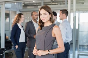 Successful formal woman standing in front of businesspeople and smiling. Portrait of confident and proud businesswoman with business team at modern office. Beautiful satisfied businesswoman looking at camera.